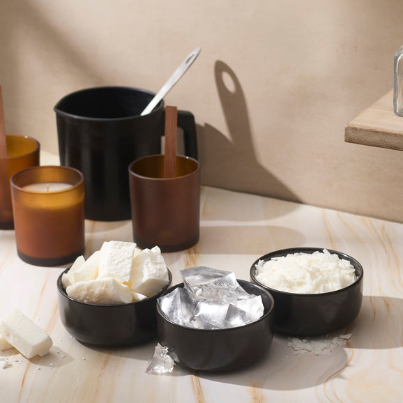 Amber glass candles and black ceramic bowls filled with wax chunks, flakes, and foil sit on a marble surface beside a black pouring pot, capturing the cozy, creative setup of a candle-making workspace.