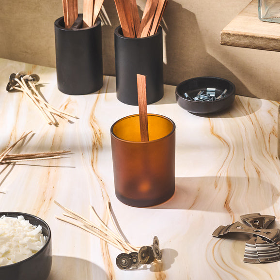 A candle-making setup featuring amber and black glass vessels, wooden wicks, wick tabs, and tools arranged on a marbled surface, capturing a warm and creative workspace moment.
