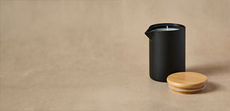 Matte black candle vessel with white wax and a bamboo lid on a neutral beige background.