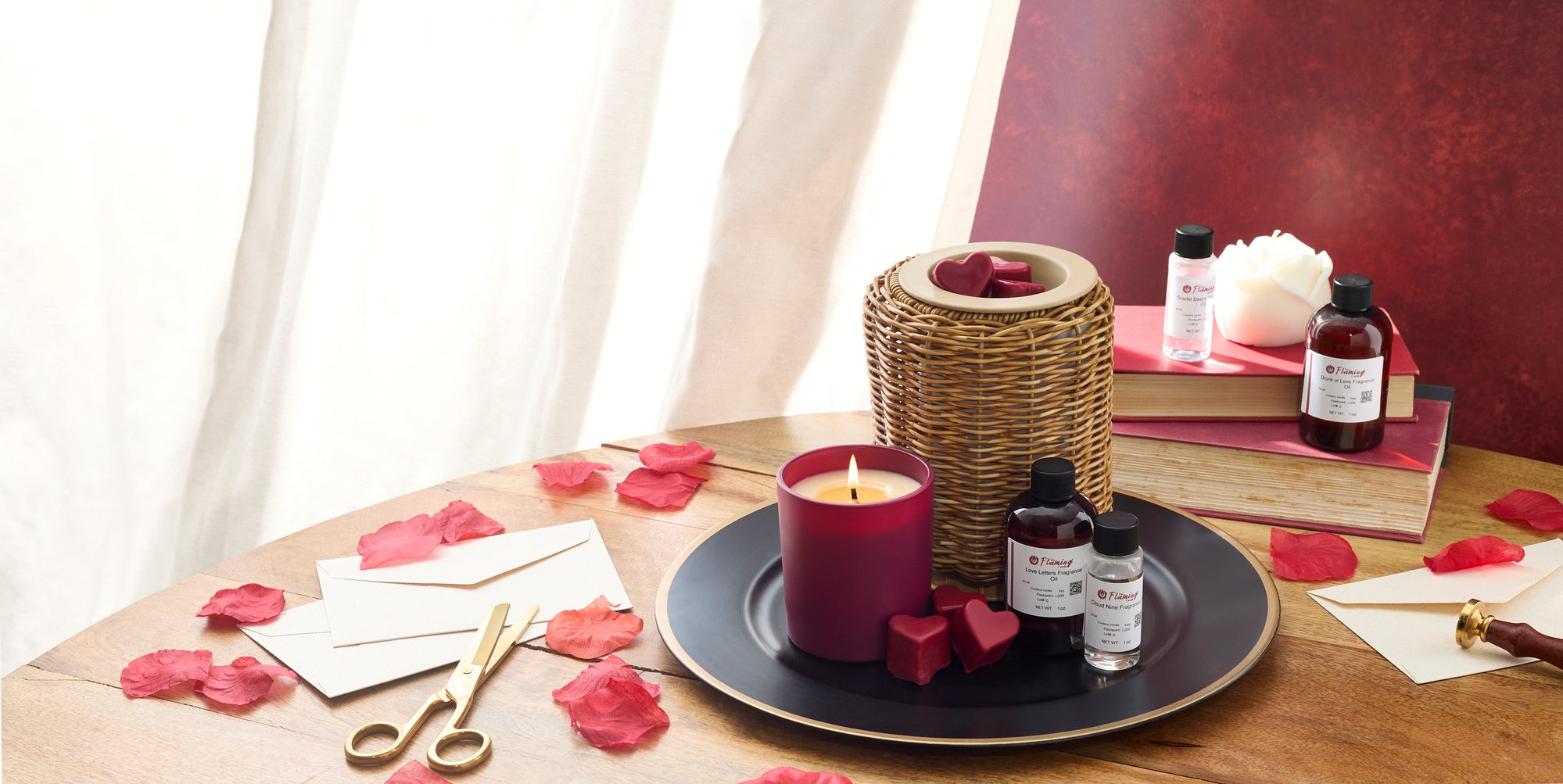 Romantic candle-making scene with a lit red candle, heart-shaped wax melts, fragrance oil bottles, rose petals, envelopes, and gold scissors styled on a wooden table with books and soft light in the background.