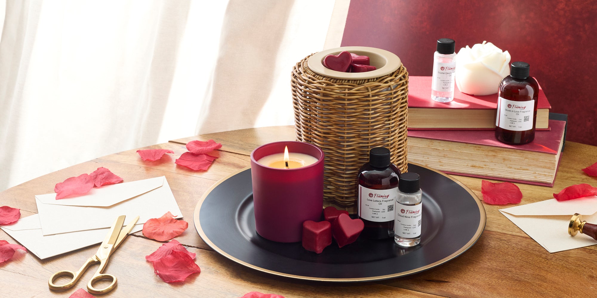 Romantic candle-making scene with a lit red candle, heart-shaped wax melts, fragrance oil bottles, rose petals, envelopes, and gold scissors styled on a wooden table with books and soft light in the background.