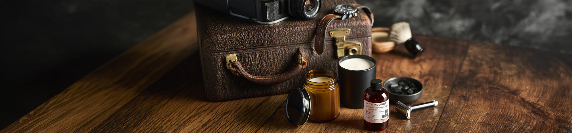 Vintage leather travel case with camera, shaving brush, razor, candle, and grooming products arranged on a wooden table in warm moody lighting.