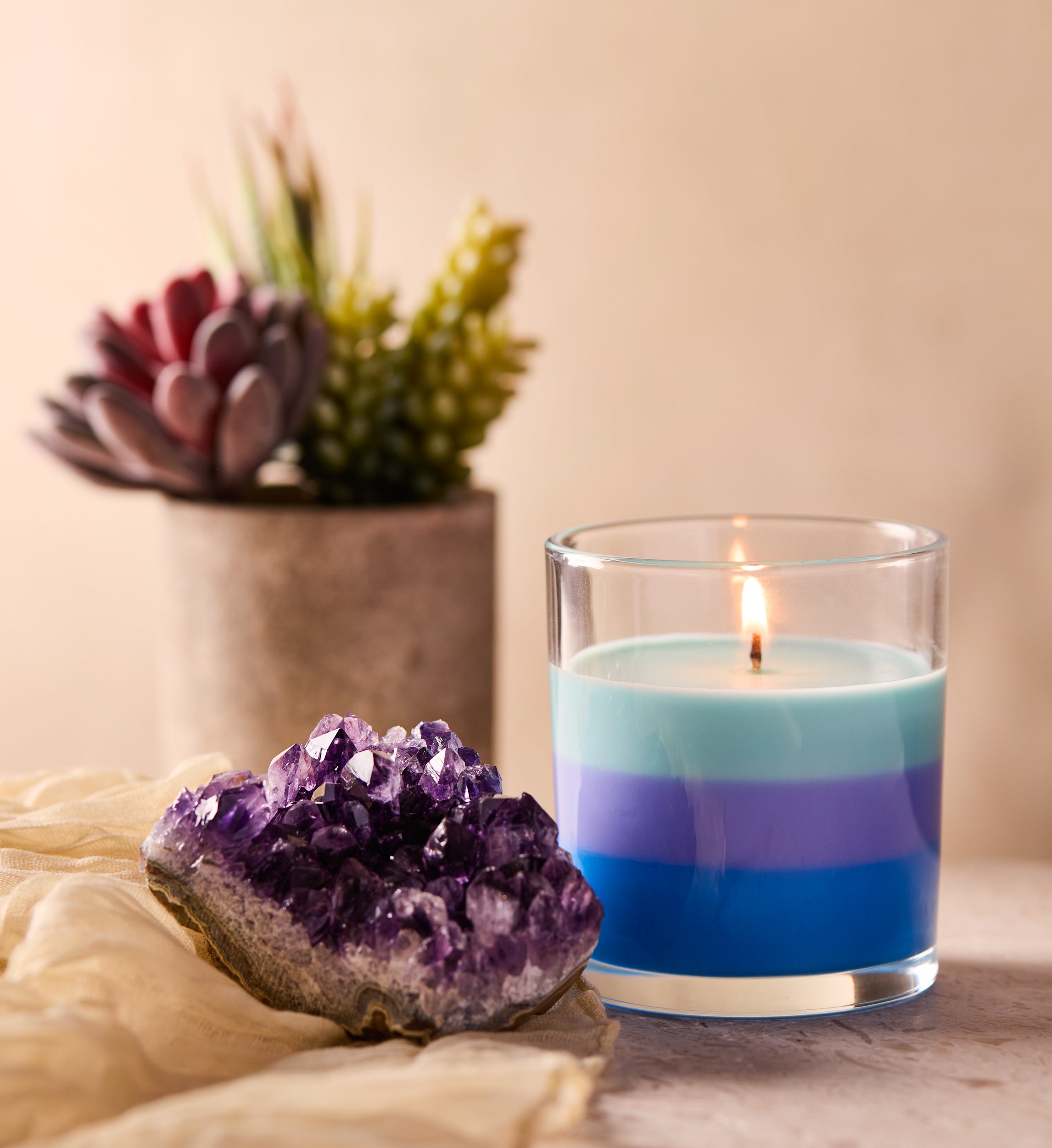 A layered blue and purple candle burning in a clear glass jar beside a cluster of amethyst crystals on a soft beige fabric.