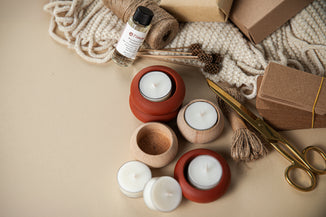 Assorted tea light candles in terracotta and wooden holders arranged on a neutral surface with twine, scissors, kraft boxes, tags, and a small bottle of fragrance oil, creating a cozy, handmade gift-making scene.