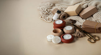 Flat lay of eco-friendly gift wrapping supplies including kraft paper boxes, brown gift tags, natural twine, brass scissors, tealight candles in wooden and terracotta holders, and a knitted cream blanket on a neutral beige background.