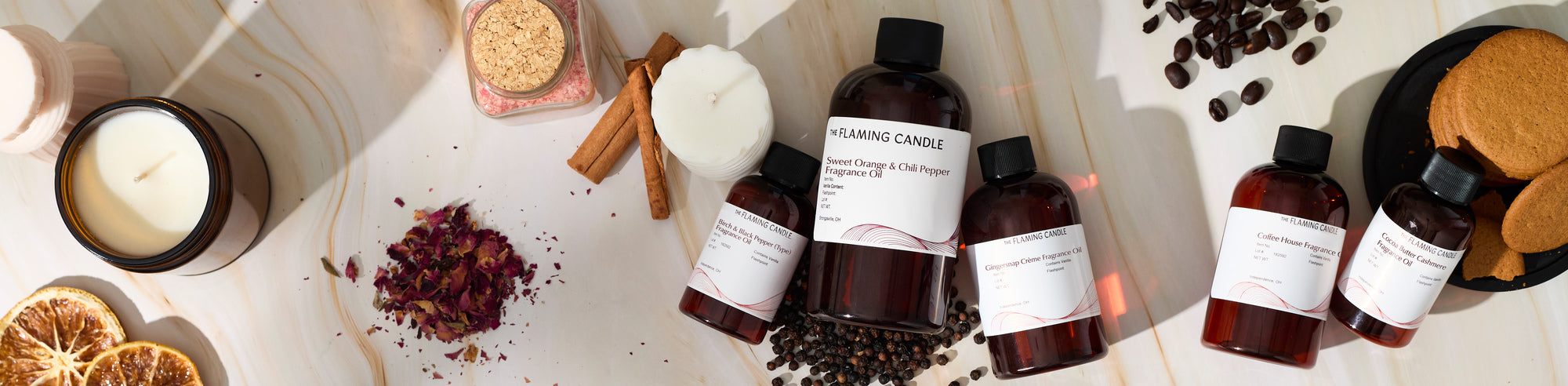 Wide flat lay of candle-making fragrance oils arranged with an amber jar candle, tealight candles, dried orange slices, cinnamon sticks, rose petals, coffee beans, peppercorns, cookies, and bath salts on a light marble surface.