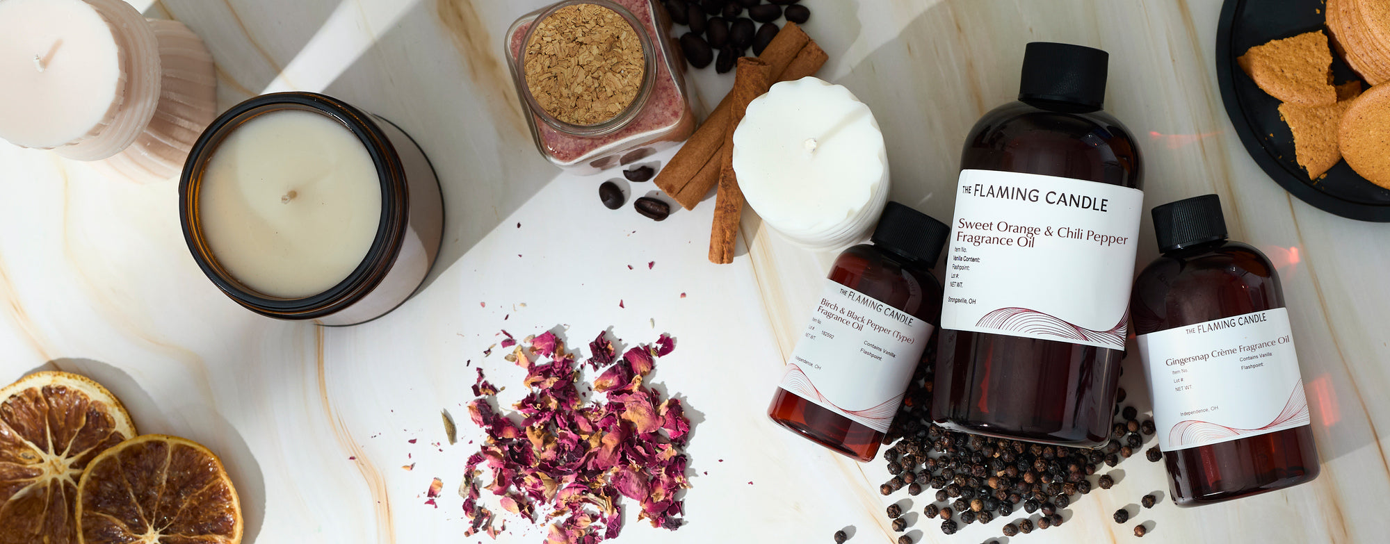 Wide flat lay of candle-making fragrance oils arranged with an amber jar candle, tealight candles, dried orange slices, cinnamon sticks, rose petals, coffee beans, peppercorns, cookies, and bath salts on a light marble surface.