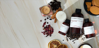 Amber fragrance oil bottles arranged with dried orange slices, coffee beans, cinnamon sticks, peppercorns, dried rose petals, and a white candle on a marble surface.