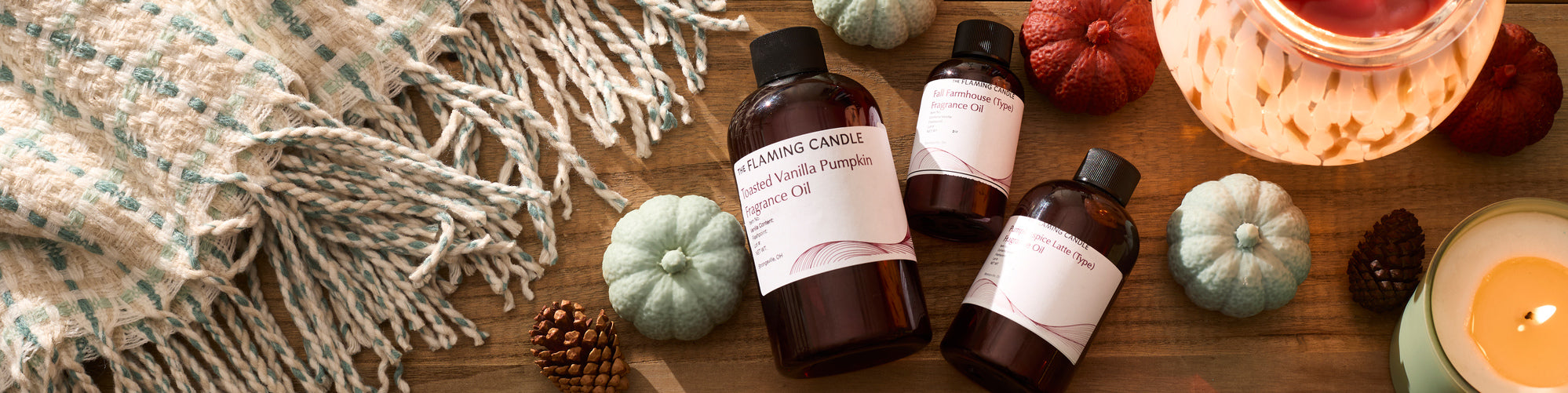 Wide flat lay of autumn-themed candle fragrance oils displayed with mini pumpkins, pinecones, a lit candle, a glowing wax warmer, and a woven cream-and-teal throw on a wooden tabletop.