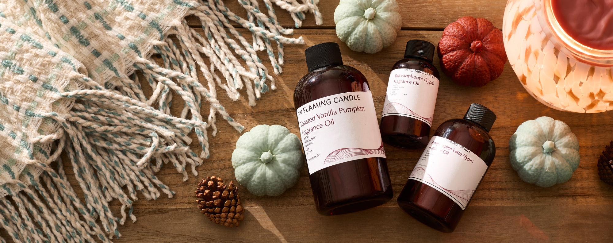 Wide flat lay of autumn-themed candle fragrance oils displayed with mini pumpkins, pinecones, a lit candle, a glowing wax warmer, and a woven cream-and-teal throw on a wooden tabletop.