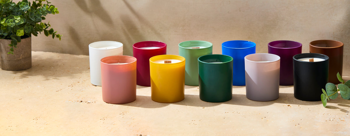 Colorful glass jar candles with wooden wicks arranged on a sunlit stone surface in white, pink, yellow, green, blue, purple, brown, and black.