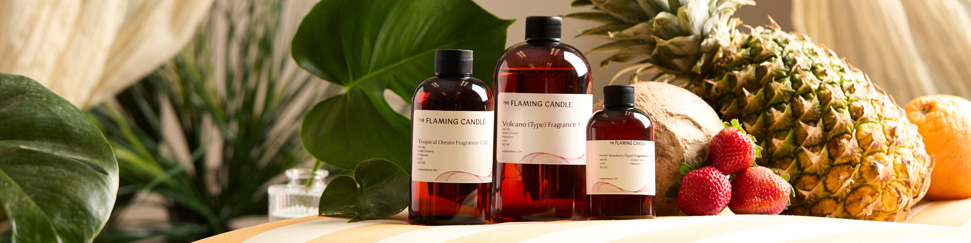 Three amber bottles of fragrance oil labeled Tropical Dream, Volcano, and Sweet Strawberry on a table with pineapple, coconut, strawberries, and tropical leaves in the background.