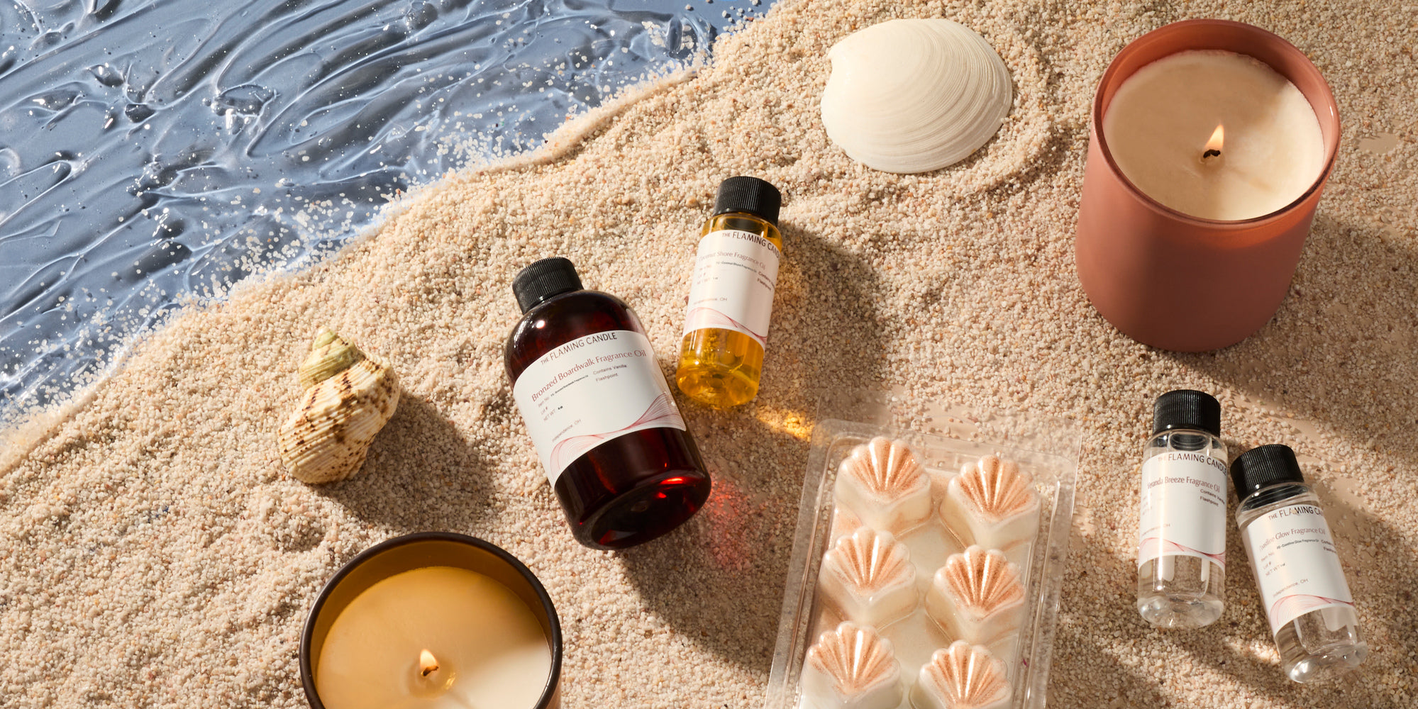 Beach-themed flat lay with lit candles, fragrance oil bottles, seashells and wax melts arranged on sand beside blue ocean waves.