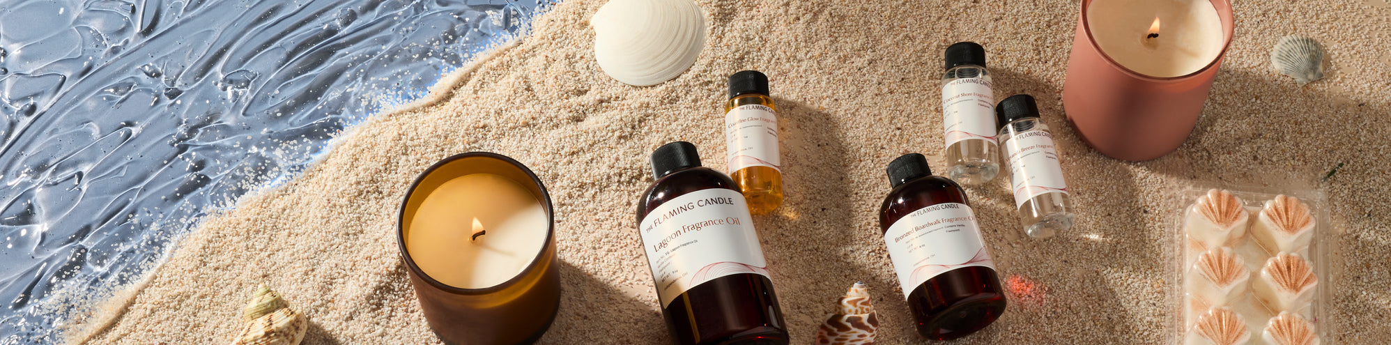Beach-themed candle and fragrance display on sand with seashells and ocean water: lit candles, bottles of Flaming Candle fragrance oil, and shell-shaped wax melts arranged in a coastal flat lay.