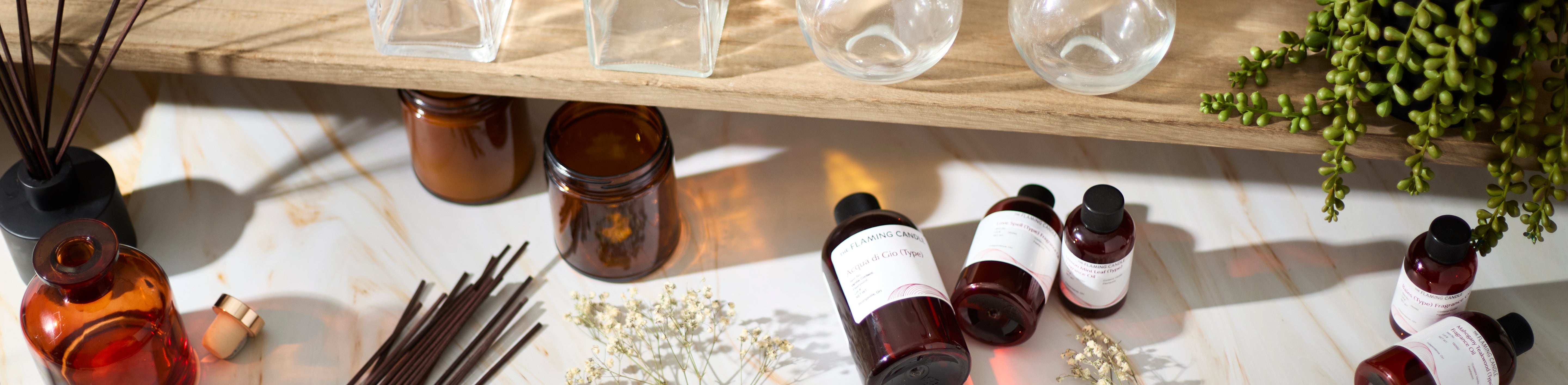 Amber glass bottles and jars, fragrance oil containers, reed diffuser sticks, dried white flowers, and hanging greenery arranged on a marble surface beneath a wooden shelf with glass vases in warm natural light.