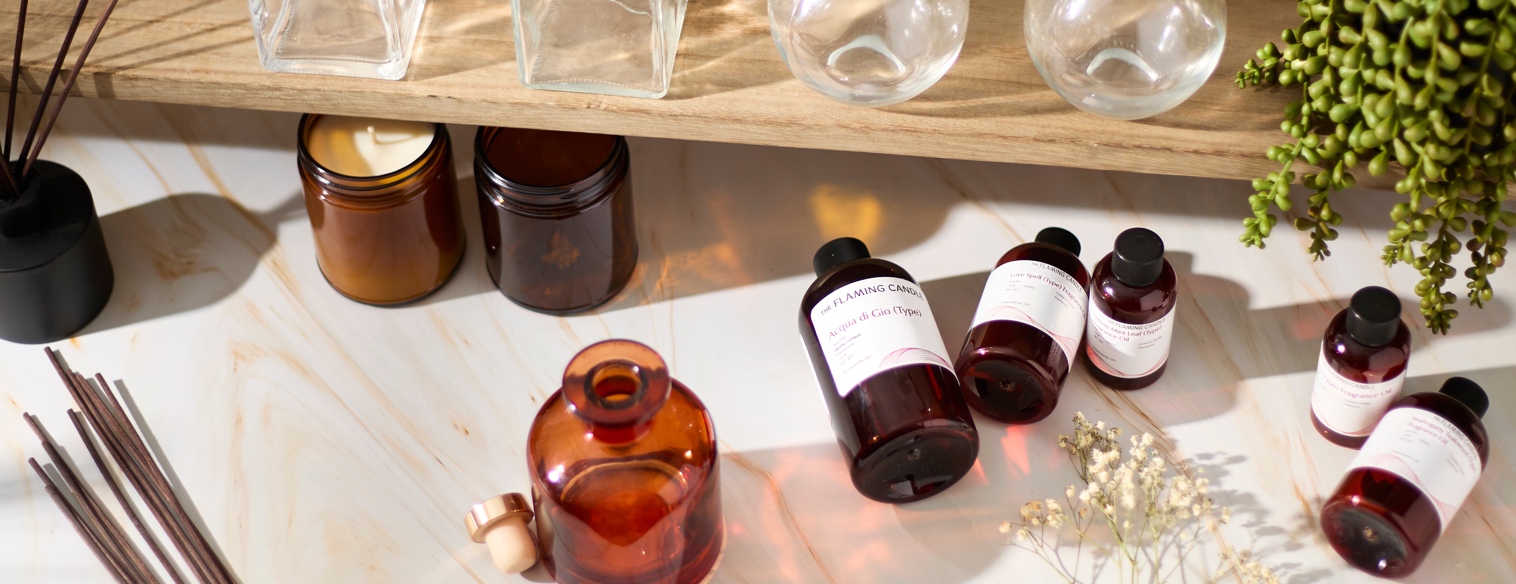 Amber glass bottles and jars, fragrance oil containers, reed diffuser sticks, dried white flowers, and hanging greenery arranged on a marble surface beneath a wooden shelf with glass vases in warm natural light.