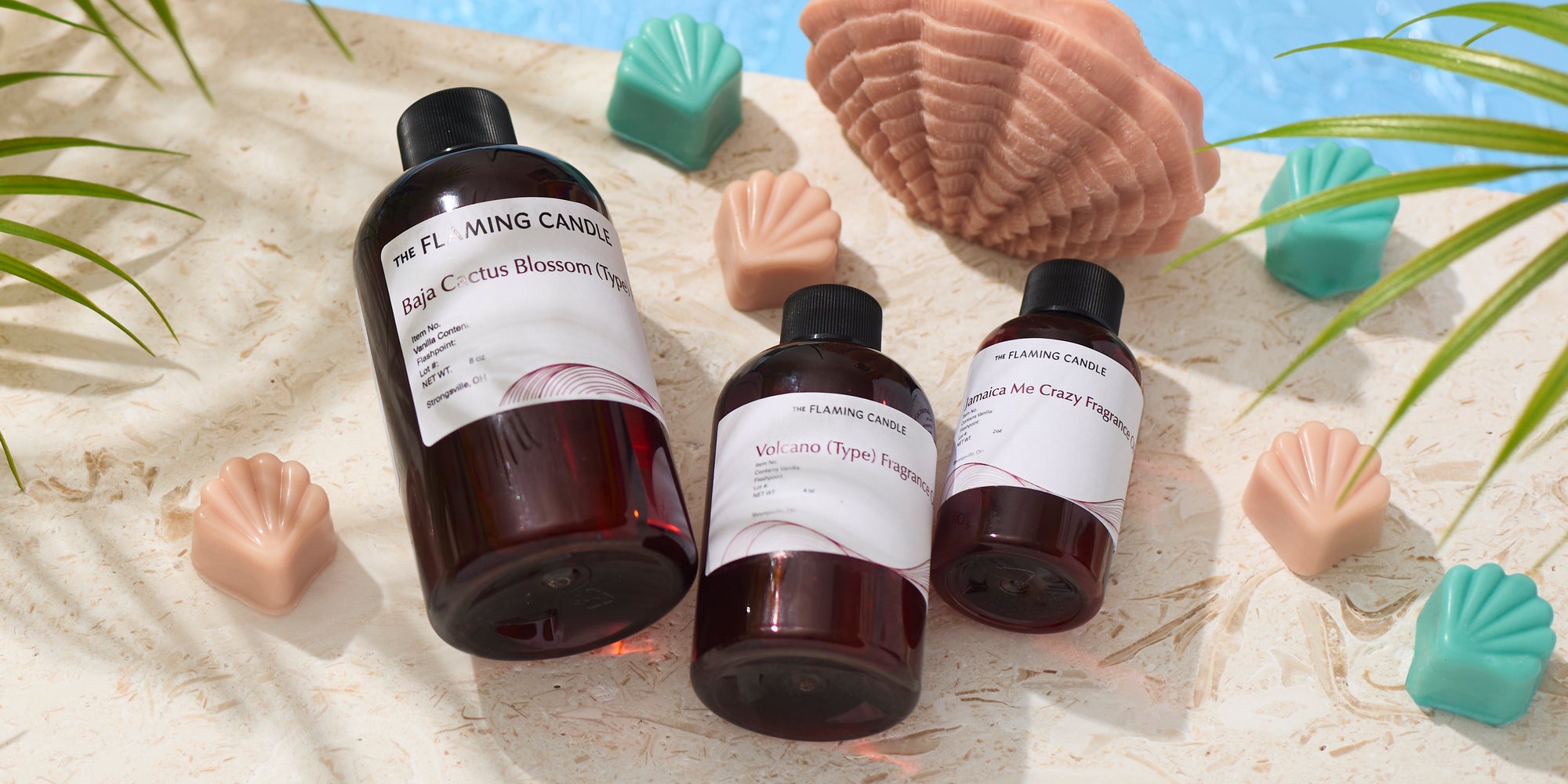 Amber bottles of fragrance oil labeled Baja Cactus Blossom, Volcano, and Jamaica Me Crazy arranged on a light stone surface with pastel shell-shaped wax melts, tropical leaves, a large seashell, and a poolside backdrop.