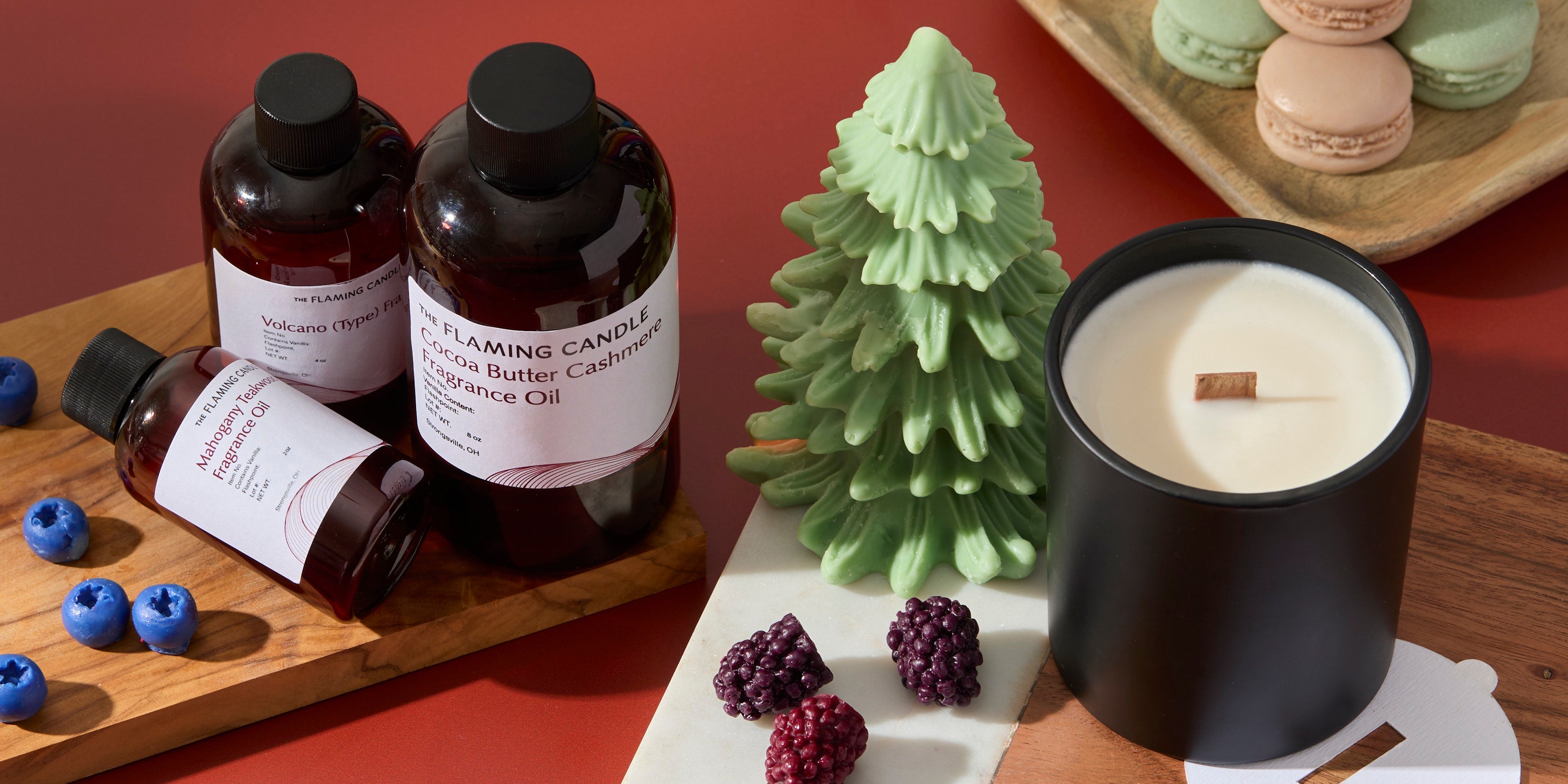 Amber fragrance oil bottles, a green tree-shaped candle, and a black wood-wick candle arranged with berries and pastel macarons on a warm red backdrop.