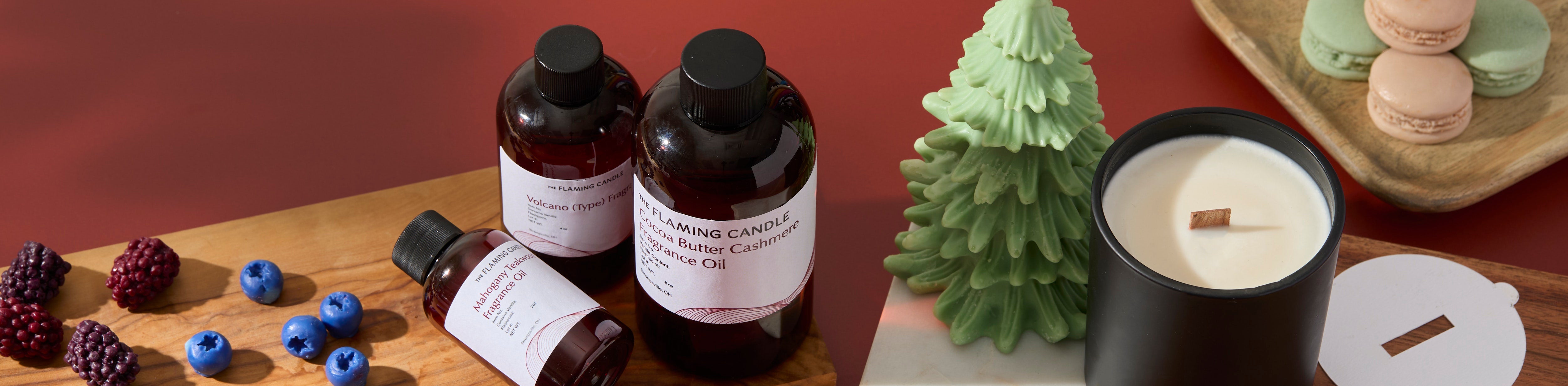 Amber fragrance oil bottles, a green tree-shaped candle, and a black wood-wick candle arranged with berries and pastel macarons on a warm red backdrop.